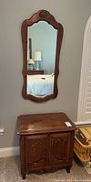 View of wooden nightstand below a mounted decorative wooden-framed mirror.