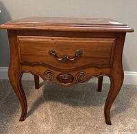 Front view of one Ethan Allen nightstand showing curved legs, carved wood decorations, and single drawer with decorative metal handle.
