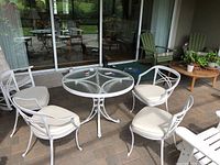 Four white cast aluminum patio chairs with beige cushions around a matching round glass-top patio table on a paved patio area.