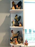 White corner shelves displaying the full set of 12 rooster and chicken figurines decorated in multi colors including black, white, beige, red and slight blue accents. Different sizes and shapes, some sitting and some standing.