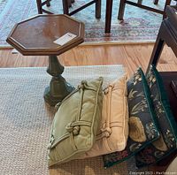 Side table and four decorative pillows arranged on a beige rug; the side table has an octagonal wooden top and green pedestal base; pillows are in green, beige, and floral designs.