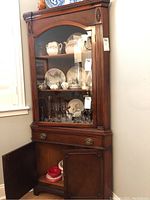 Full view of vintage cherry wood corner buffet curio cabinet with glass front upper display filled with dishware, one drawer and open lower cabinet door showing inside storage.
