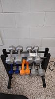 Wide-angle photo showing six hex dumbbells resting on a black metal rack. The visible weights in this photo are 5 lb, 8 lb, 10 lb, and 15 lb. Also includes a pair of orange neoprene dumbbells and blue dumbbells. The dumbbell rack holds two rows of weights.