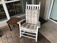 Overall view of white rocking chair with beige cushions and adjacent round wooden side table on patio floor.