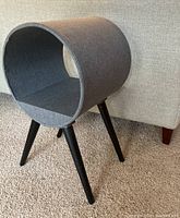 Side view of a modern gray fabric covered circular cat bed with black angled legs on carpet.