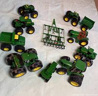 Top-down view of the nine John Deere green and yellow toy tractors arranged around a green tillage implement with multiple tines on a light surface.