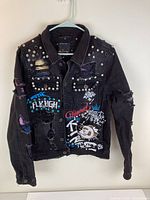 Front view of black denim jacket showing studs on shoulders, distressed fabric, colorful prints and patches on front.
