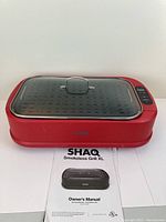Front view of red SHAQ Smokeless Grill XL with transparent lid and handle on top. Owner's manual visible beneath the grill.