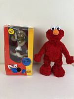 Singing Homer Simpson figure in original box with visible wear next to a red Elmo plush toy against a plain background.