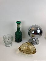 All four display items including a green glass decanter, clear embossed glass goblet, round chrome incense burner, and amber tinted etched glass ashtray.