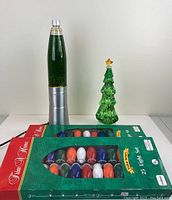 Photo showing green glitter lava lamp missing top, green glass Christmas tree figurine, and two boxes of vintage 25 multicolor outdoor Christmas lights.
