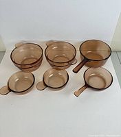 Top-down and side view of 6 amber tinted glass mixing bowls and measuring cups with integrated handles arranged in two rows on white background.
