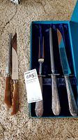 Three Birks sterling silver carving utensils in velvet-lined blue box beside Premier stainless steel carving knife and sharpener on carpeted floor
