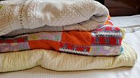 Three blankets stacked on a bed, clearly showing the beige crocheted blanket on top, the colorful patchwork blanket in the middle, and the yellow quilted blanket at the bottom.