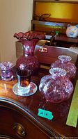 Photo showing cranberry glass items including a 6-inch vase, two small vases, a candle holder on mirror tray, and two ruby overlay cologne bottles on wood surface.