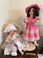 Two vintage dolls, one standing in a pink lace dress, the other seated in a white lace dress holding a parasol and teddy bear.