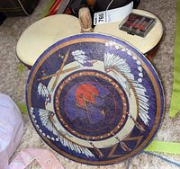 Photo of signed indigenous style hand drum showing feather-arrow painted design and surface wear