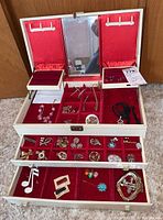 Open cream jewelry box with red velvet interior. Mirror center with compartments holding various costume jewelry including earrings and necklaces.
