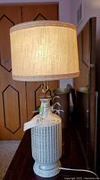 Full view of vintage ceramic base lamp with beige fabric drum shade on top of a wooden surface.