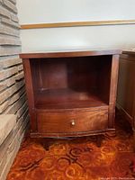 Front view of the wood side table showing open shelf and drawer closed, positioned next to stone wall with patterned carpet below.