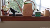 Overview of all items: plant, black bird figurine, pink ceramic watering can, smaller beige watering can, Schultz liquid plant food bottle on windowsill.