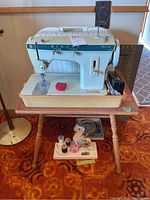 Singer Fashion Mate 334 sewing machine on cream base with red spool holder and foot pedal visible on side. Machine set on wooden table with metal legs. Accessories including thread spools below the table.