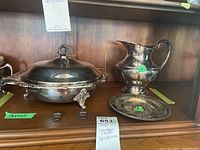 Photo of the silver plated serving dish with ornate handles and claw feet, water jug, and small round tray on a wooden shelf.