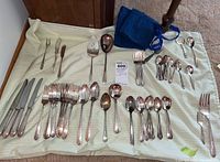 Full view of assortment of silver plate cutlery laid on fabric including forks, knives, spoons, serving spoons, with two felt storage wraps.