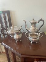 Full view of the silver plate tea set consisting of four pieces on a wooden surface: tea pot, coffee pot, creamer, and sugar bowl, all with ornate silver plating and wooden handles on the larger pots.