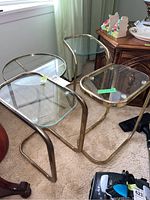 Set of four glass and metal side tables shown together. Metal frames tarnished and some rust visible. Different shapes and sizes, metal tubular construction, clear glass tops.