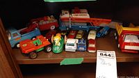 Shelf containing various vintage toy cars and trucks in used condition with scratches and signs of wear. Vehicles include pickup trucks, transport trucks, small cars, and novelty toys in assorted colors.