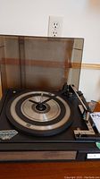 Front view of Sears turntable with transparent dust cover in place, black platter with silver-grey detailing, black tonearm, and wooden front panel. Power outlet in background.