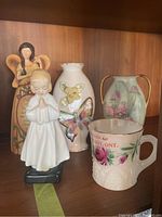 Photo showing all five items together on a shelf: two vases, praying child figurine, angel figurine, and souvenir cup.