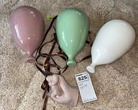 Group photo of three ceramic balloons in pink, green, white along with a ceramic hand holding brown ribbons, placed on carpet.