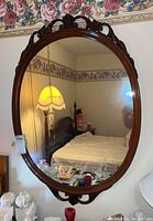 Front view of the oval mirror hanging on a wall in a bedroom, showing ornate carved wood frame and reflective surface.