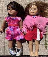 Two 18-inch American Girl dolls displayed outdoors on paved ground. Luciana doll on left, darker skin tone, galaxy dress, holographic boots. Truly Me doll on right, lighter skin, red and white dress, pink poncho, face marks visible.