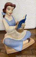 Full view of the Bookish Beauty Belle figurine sitting cross-legged with blue dress, white apron, holding blue book and basket on side.