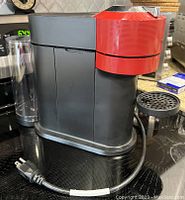 Side view of the coffee machine showing red upper portion with textured finish, black body, water reservoir, and power cord.