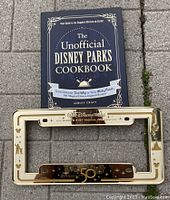 Photo showing The Unofficial Disney Parks Cookbook with dark blue cover and gold text, alongside a cream and gold 50th anniversary license plate cover.