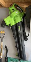 GreenWorks electric leaf blower/vacuum unit in green and black with visible power cord and handle.