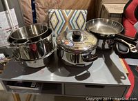Photo of stainless steel pots and pans arranged on a table, showing multiple sizes including frying pan, pots with lids, and a strainer.