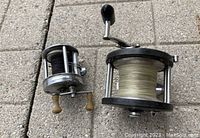 Two vintage fishing reels placed side by side on a tiled surface, one smaller with dark line and another larger with light-colored line.