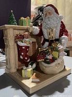 Front view showing Santa with sack of toys next to fireplace, stocking, tree, and gifts
