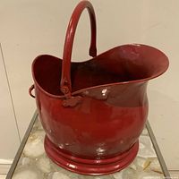 Side view of red painted antique brass coal scuttle showing the broad spout and curved handle.