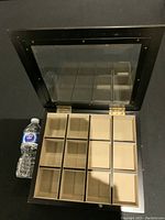 Open black watch/jewelry box showing twelve compartments lined in beige fabric, with water bottle for scale, and metal hinges on glass lid.