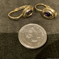 Pair of 18K yellow gold amethyst drop earrings displayed on a dark cloth with a coin for size reference