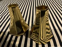 Pair of heavy, polished brass candle holders on striped fabric background, showing flared hexagonal shape and reflective surface.