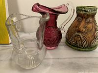 All three vintage jugs shown together on a surface: clear glass jug, purple glass jug, and pottery jug with a bearded face