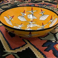 Side angle showing the yellow ceramic bowl with white and red hand-painted whirling dervish figures inside and around the exterior.