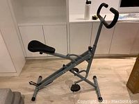 Side view of the Upright Row-N-Ride rowing machine showing padded seat, pedals and handlebar with foam grips, placed in a room with wood floor.